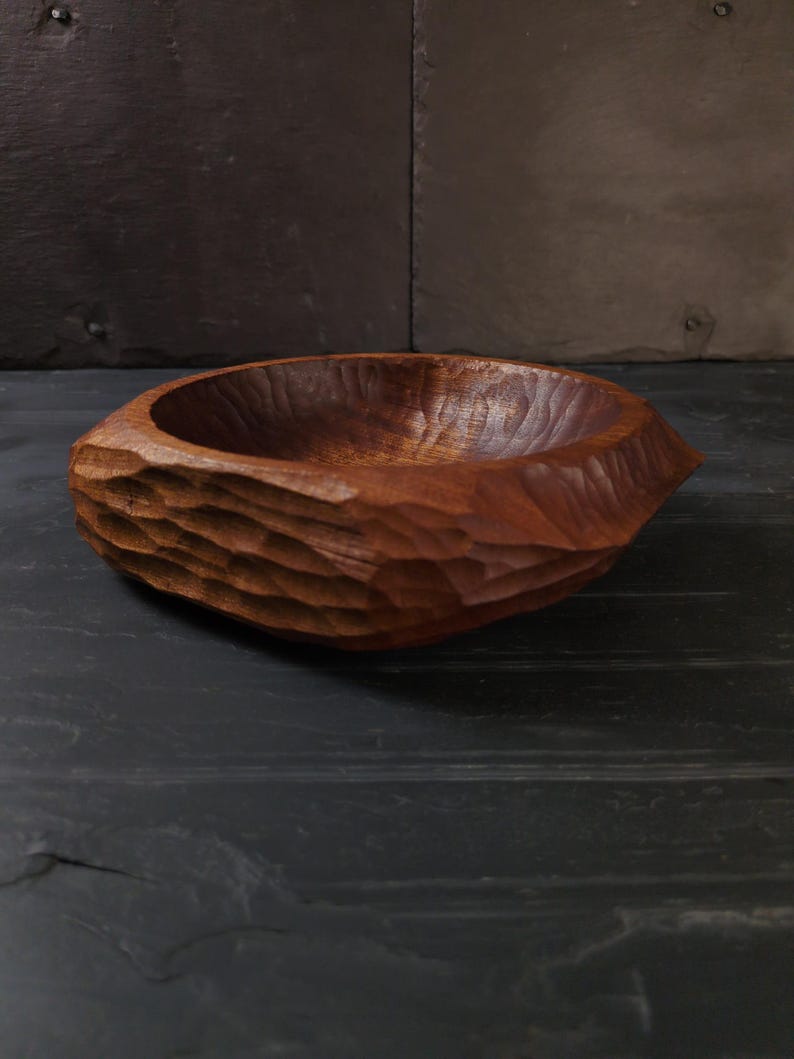 May include: A hand-carved wooden bowl with a textured exterior and a smooth interior. The bowl is a rich brown colour and appears to be made from a single piece of wood. The bowl is on a dark surface.