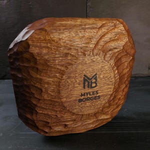 May include: A hand-carved wooden bowl with a textured, organic design. The bowl is a warm brown colour, with a lighter area on the top edge. The bottom of the bowl features the text "MYLES BORGES" and a logo.