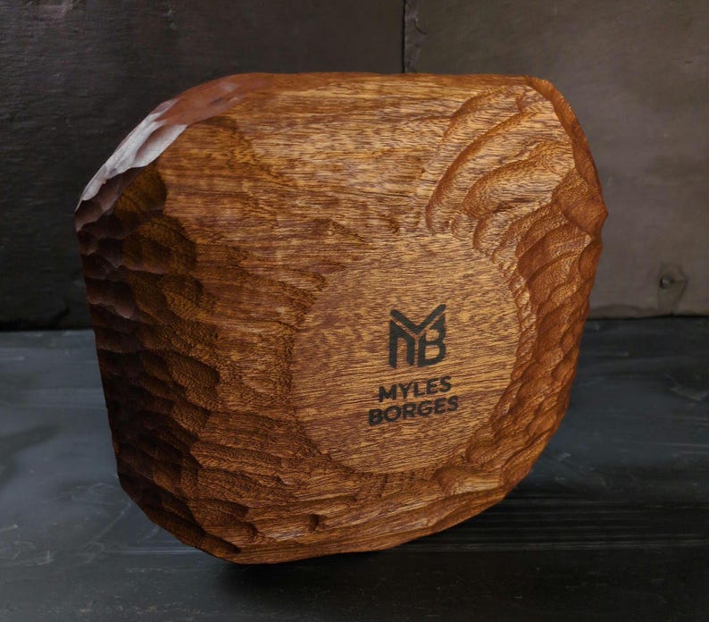 May include: A hand-carved wooden bowl with a textured, organic design. The bowl is a warm brown colour, with a lighter area on the top edge. The bottom of the bowl features the text "MYLES BORGES" and a logo.