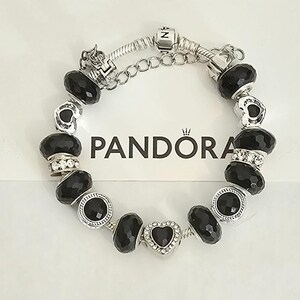 May include: A silver Pandora bracelet with black faceted beads and heart-shaped charm. The bracelet features a chain closure and is displayed on a white surface with the Pandora logo.