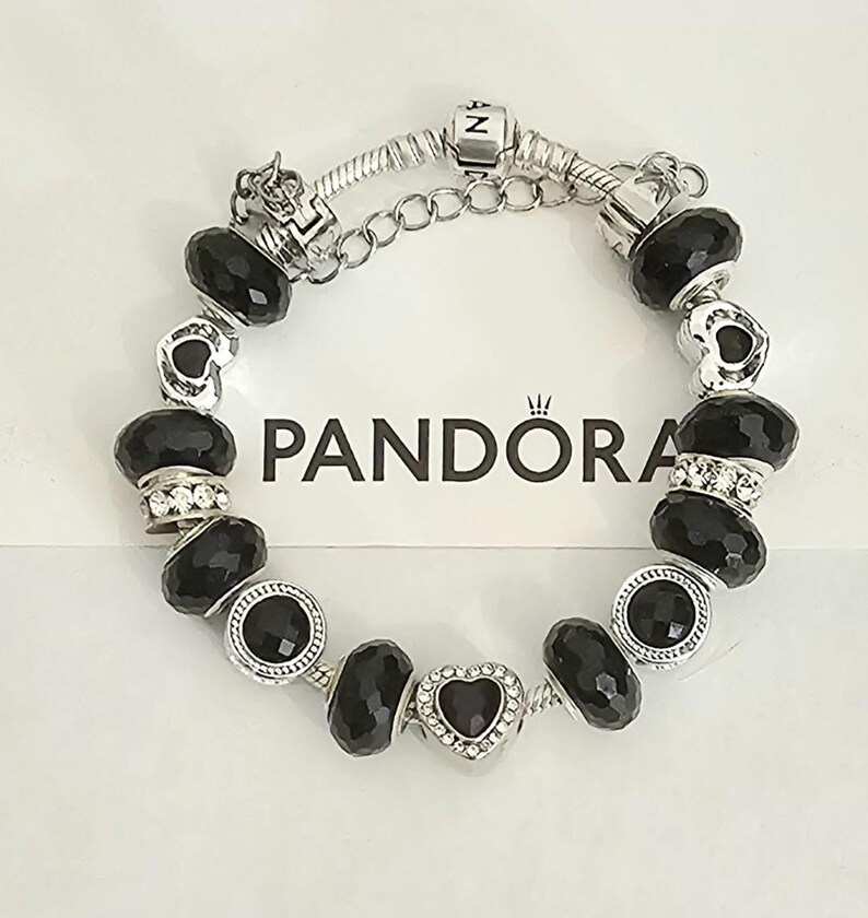 May include: A silver Pandora bracelet with black faceted beads and heart-shaped charm. The bracelet features a chain closure and is displayed on a white surface with the Pandora logo.