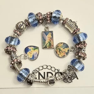 May include: A silver charm bracelet featuring light blue faceted glass beads, pink crystal-studded spacers, and three dangling charms depicting a fairy character. The bracelet includes a metal clasp and a chain extender with a silver-toned finish.