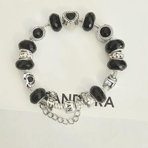 May include: A silver charm bracelet with black faceted beads and decorative charms. The bracelet includes a heart-shaped charm and charms with the letters "PAN". The bracelet is on a white surface.
