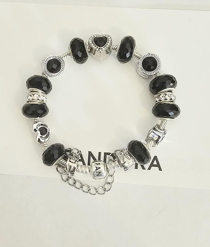 May include: A silver charm bracelet with black faceted beads and decorative charms. The bracelet includes a heart-shaped charm and charms with the letters "PAN". The bracelet is on a white surface.