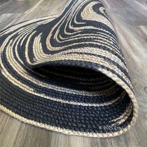 May include: A rolled-up, oval-shaped rug with a black and beige swirl pattern. The rug is made of woven material and has a braided border. The rug's design creates a wave-like effect.