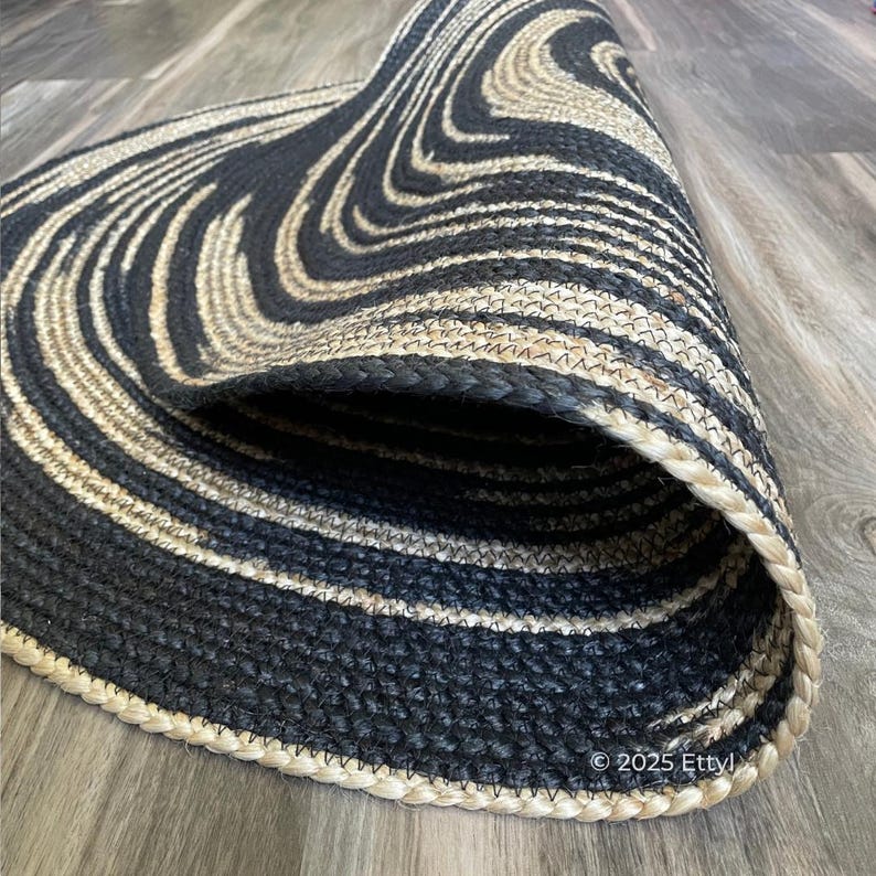 May include: A rolled-up, oval-shaped rug with a black and beige swirl pattern. The rug is made of woven material and has a braided border. The rug's design creates a wave-like effect.