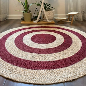 May include: A round jute rug with a concentric design. The rug features alternating bands of natural beige and deep red. The rug is placed on a dark wood floor, with plants and a small table in the background.