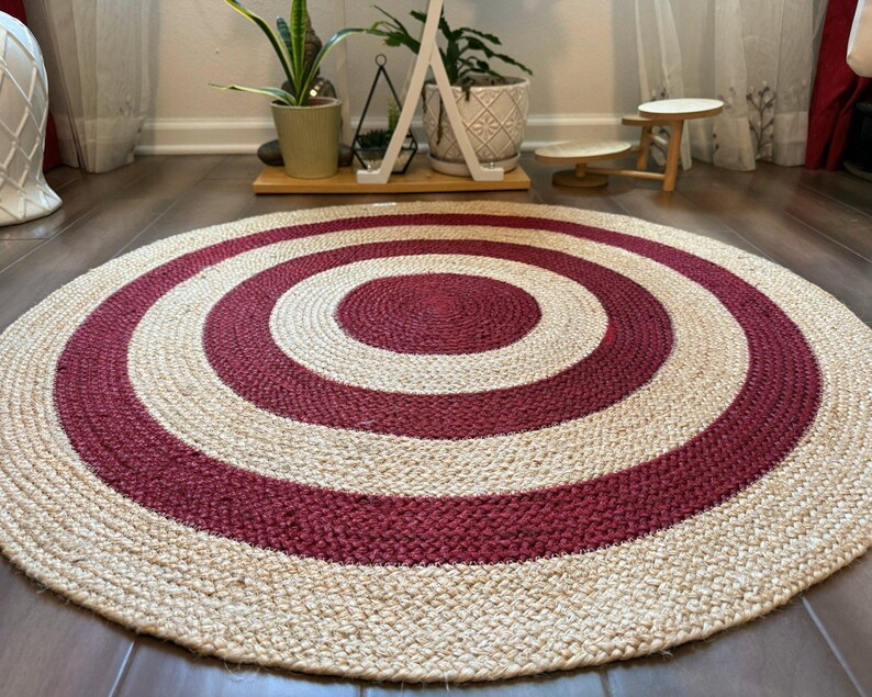 May include: A round jute rug with a concentric design. The rug features alternating bands of natural beige and deep red. The rug is placed on a dark wood floor, with plants and a small table in the background.