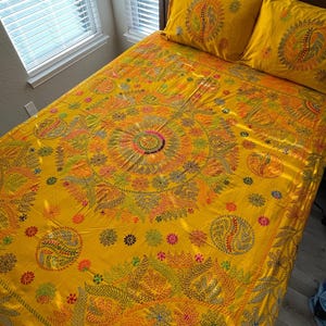 May include: A yellow bedspread and two matching pillowcases with intricate, colorful embroidered floral and paisley designs. The bedding is on a wooden bed frame, creating a warm and inviting bedroom scene.