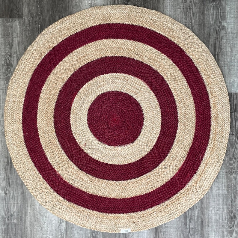 May include: A round jute rug with a concentric design. The rug features alternating bands of natural beige and deep red. The central circle and the bands are a deep red color. The rug is made of woven jute.
