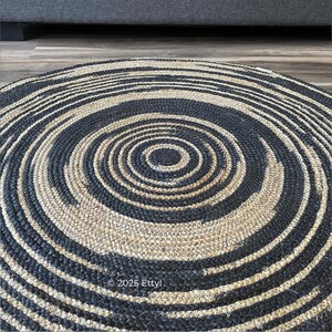 May include: A round rug with a spiral design. The rug features alternating bands of black and natural beige fibers, creating a concentric pattern. The rug is made of woven material and has a textured surface. The text "&copy;2025 EttyI" is visible.