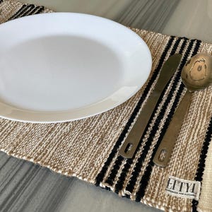 May include: A white oval plate rests on a beige and black striped woven placemat, with a knife and spoon to the side. The placemat has a tag that reads "EITYL HANDMADE FOR ELEGANCE".