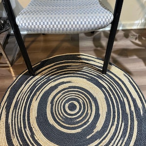 May include: A round rug with a spiral design in black and beige. The rug is made of woven material and has a concentric pattern. The rug is placed under a chair with a checkered seat.