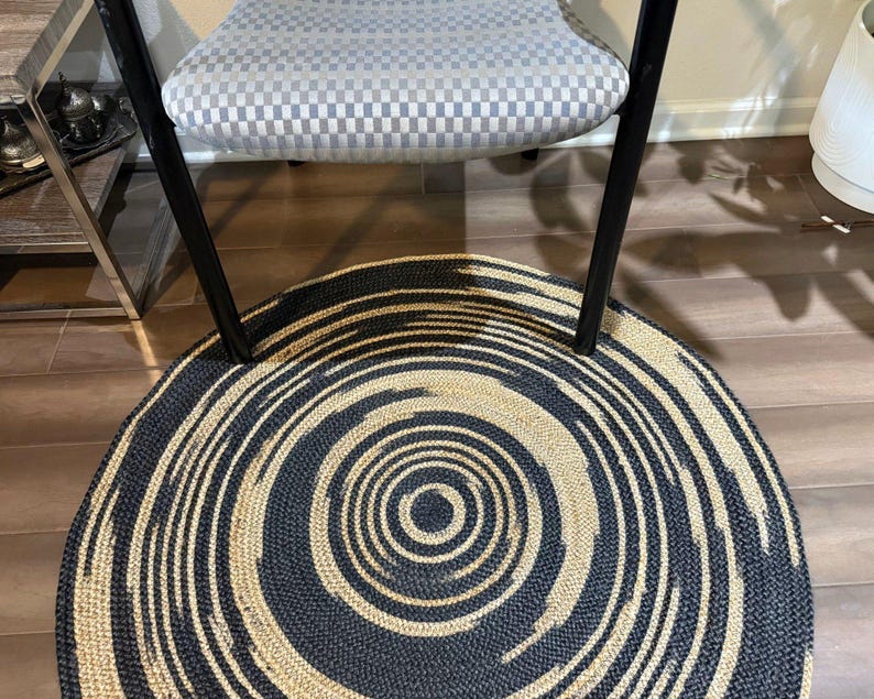 May include: A round rug with a spiral design in black and beige. The rug is made of woven material and has a concentric pattern. The rug is placed under a chair with a checkered seat.