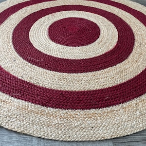 May include: A round jute rug with a concentric design. Alternating bands of natural beige and deep red create a target-like pattern. The rug's texture is visible, showcasing the woven fibers. This rug is suitable for living rooms and bedrooms.