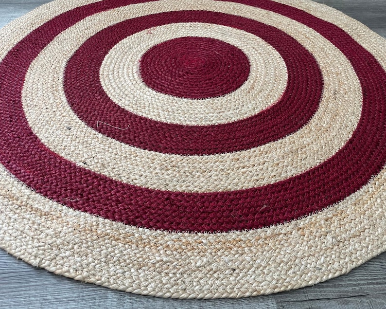 May include: A round jute rug with a concentric design. Alternating bands of natural beige and deep red create a target-like pattern. The rug's texture is visible, showcasing the woven fibers. This rug is suitable for living rooms and bedrooms.