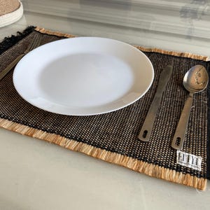 Puede incluir: Un plato blanco, tenedor, cuchillo y cuchara sobre un mantel individual tejido negro y beige con flecos. El mantel individual tiene forma rectangular y está colocado sobre una superficie clara. La cubertería es de acero inoxidable.