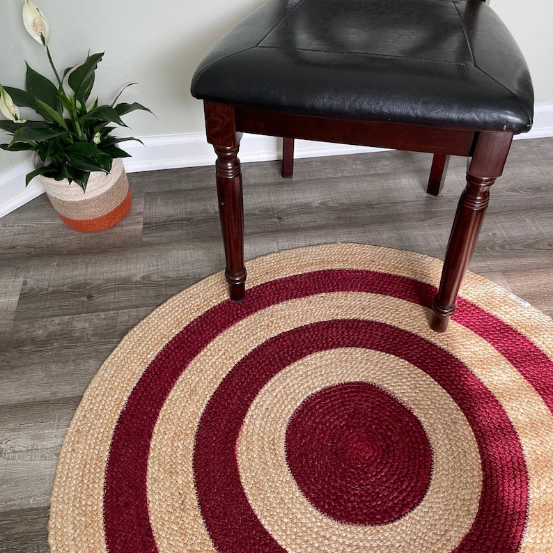 May include: A round jute rug with concentric circles in natural tan and burgundy. The rug is placed on a wooden floor, with a chair and a potted plant in the background.