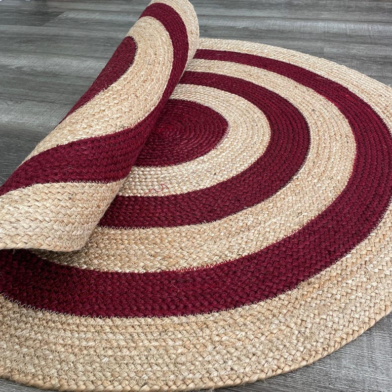 May include: A round jute rug with a spiral design. The rug features alternating bands of burgundy and natural beige. The rug is partially rolled up, revealing the underside and the texture of the woven material.