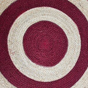 May include: Circular jute rug with a bold design. The rug features concentric circles in burgundy and natural beige. The texture is visible, showcasing the woven construction. This rug is suitable for various interior decor styles.
