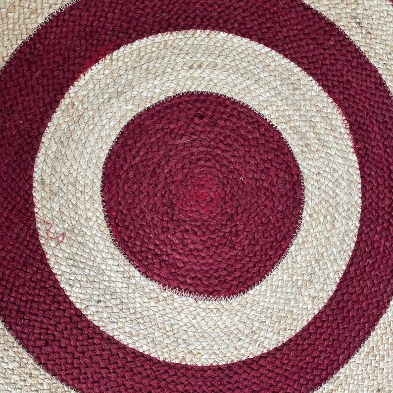 May include: Circular jute rug with a bold design. The rug features concentric circles in burgundy and natural beige. The texture is visible, showcasing the woven construction. This rug is suitable for various interior decor styles.