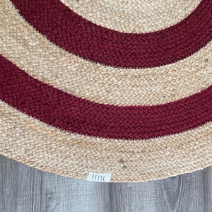 May include: A round jute rug with alternating bands of natural beige and deep red. The rug has a woven texture and a label with the text "FITYL" is visible in the lower corner. This rug is suitable for indoor use.