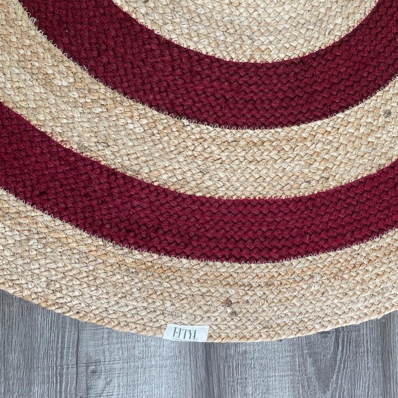 May include: A round jute rug with alternating bands of natural beige and deep red. The rug has a woven texture and a label with the text "FITYL" is visible in the lower corner. This rug is suitable for indoor use.
