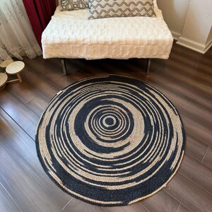 May include: A round rug with a spiral design. The rug features alternating bands of black and natural beige, creating a concentric pattern. The rug is made of woven material and is placed on a hardwood floor.