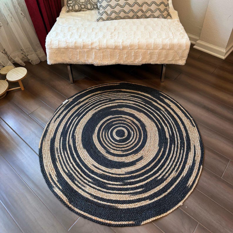 May include: A round rug with a spiral design. The rug features alternating bands of black and natural beige, creating a concentric pattern. The rug is made of woven material and is placed on a hardwood floor.