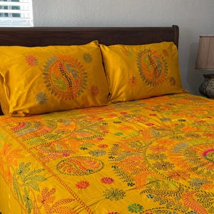 May include: A bed set featuring a yellow comforter and two pillowcases. The bedding has an intricate embroidered design in shades of orange, green, and blue. The pattern includes floral and paisley motifs, creating a vibrant and decorative look.