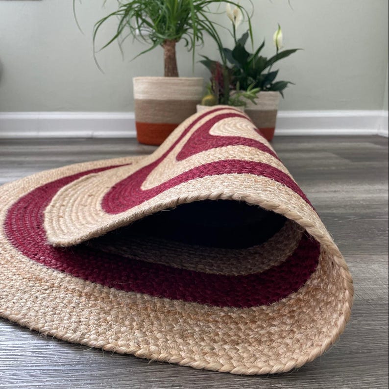 May include: A rolled-up, oval-shaped jute rug with a burgundy wave pattern. The rug is made of natural fibers and has a neutral color palette. The rug is on a gray wooden floor, with potted plants in the background.