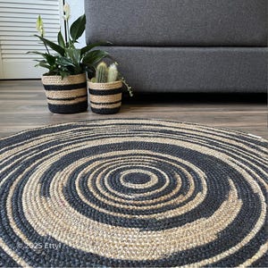May include: A round rug with a spiral design in black and natural jute. The rug is on a wooden floor, with two potted plants in black and natural striped pots and a gray sofa in the background.