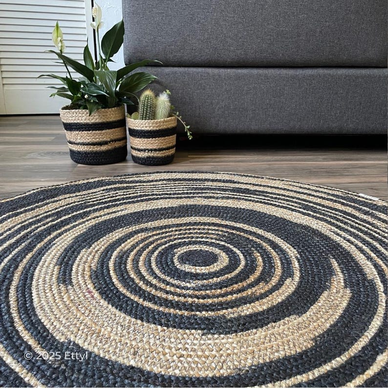 May include: A round rug with a spiral design in black and natural jute. The rug is on a wooden floor, with two potted plants in black and natural striped pots and a gray sofa in the background.