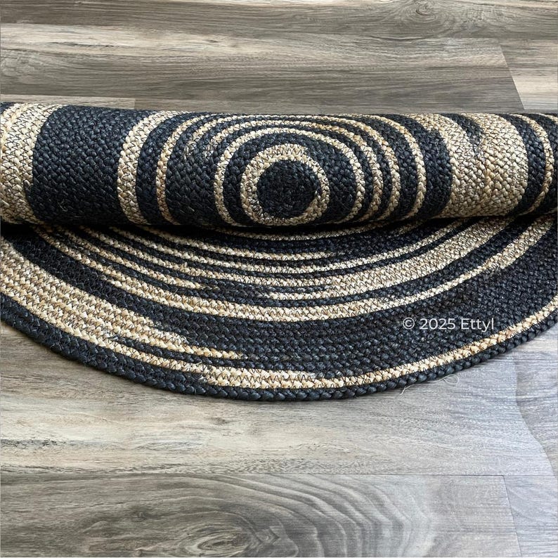May include: A rolled-up, circular jute rug with a concentric design of alternating black and natural beige bands. The rug is made of woven fibers and has a textured appearance. The rug is on a wooden floor.