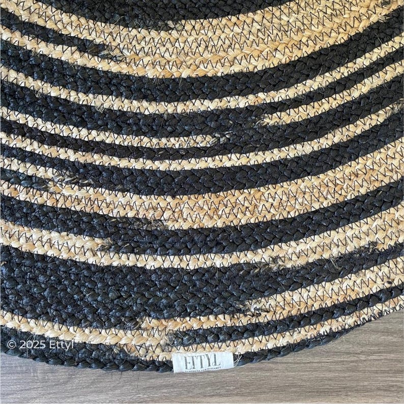 May include: A round rug with alternating black and natural-colored stripes. The rug is made of woven material and has a textured surface. The brand name "Ettyl" is visible on a small tag.