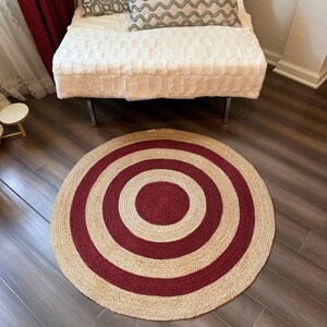 May include: A round jute rug with a target design. The rug features alternating bands of natural beige and deep red, creating a bold, concentric pattern. The rug is placed on a dark wood floor.