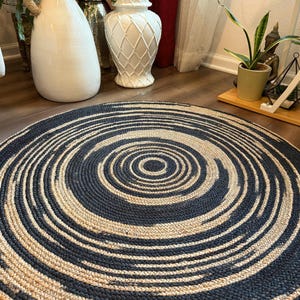 May include: A round rug with a concentric spiral design. The rug features alternating bands of navy blue and natural beige, creating a textured, woven appearance. The rug is placed on a wooden floor, with decorative vases and plants in the background.