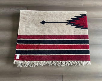 May include: A rectangular woven rug with a beige background and a geometric design in red, black, and white. The rug features horizontal stripes and a fringed edge. A black diamond and arrow design is on the top.