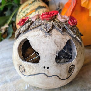 Könnte beinhalten: Eine runde, cremeweiße Keramikskulptur mit abgenutzter Oberfläche, die zwei große, hohle Augenhöhlen und einen dünnen, gewellten Mund aufweist. Die Oberseite ist mit einer Krone aus geformten dunkelgrünen Blättern und kleinen roten Rosen verziert.