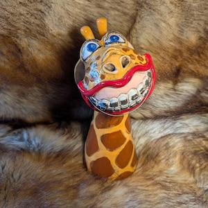 Könnte beinhalten: Eine Giraffenfigur mit einem beigen und braunen Fleckenmuster, zwei gelben Hornzapfen und großen blauen Augen. Die Giraffe hat einen breiten, lächelnden Mund mit leuchtend roten Lippen und sichtbaren silberfarbenen Zahnspangen aus Metall.