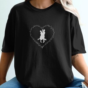 May include: Black t-shirt featuring a white heart-shaped design with a silhouette of two people dancing. The design is surrounded by small dots. The t-shirt is made of a soft material.