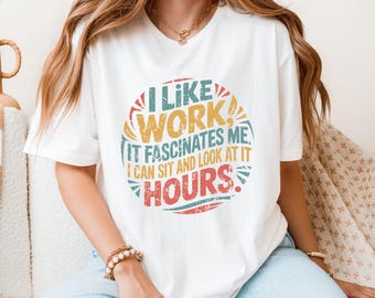 May include: White t-shirt with a colorful graphic that reads "I LIKE WORK, IT FASCINATES ME, I CAN SIT AND LOOK AT IT HOURS." The design features a circular shape with text in blue, yellow, red, and green. The shirt is a casual wear item.