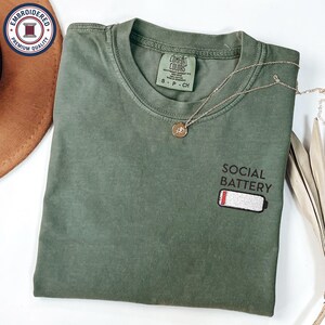 May include: Olive green t-shirt with the words "SOCIAL BATTERY" and a low battery graphic embroidered on the front. A gold necklace with a pendant is draped across the shirt. The shirt is size S-P-CH.