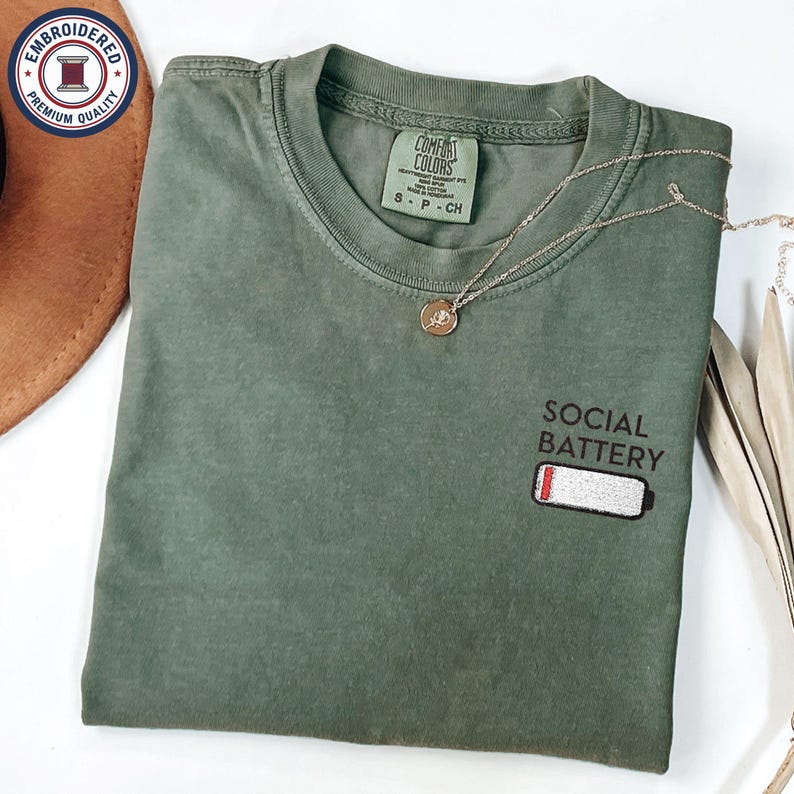 May include: Olive green t-shirt with the words "SOCIAL BATTERY" and a low battery graphic embroidered on the front. A gold necklace with a pendant is draped across the shirt. The shirt is size S-P-CH.