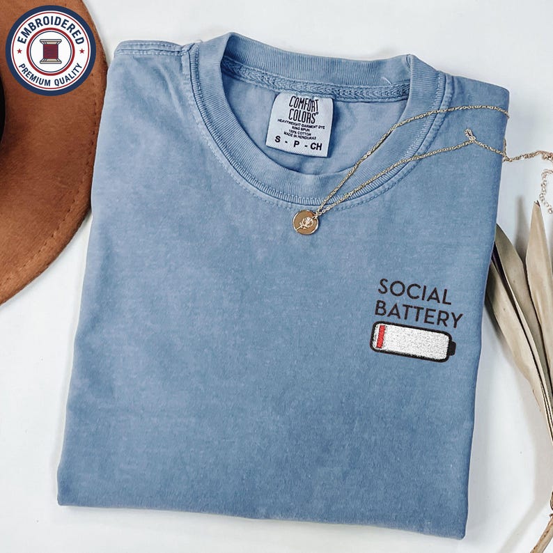 May include: A blue Comfort Colors t-shirt with the text "SOCIAL BATTERY" and a low battery graphic embroidered on the front. A gold necklace with a pendant rests on the shirt. The shirt is folded.