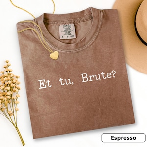 May include: Espresso-colored t-shirt with the phrase "Et tu, Brute?" in white. A gold heart pendant necklace rests on the shirt. A tan hat and a sprig of dried flowers are also visible.