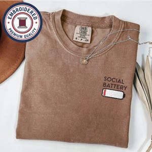 May include: A brown Comfort Colors t-shirt with the words "SOCIAL BATTERY" and a low battery graphic embroidered on the front. A silver necklace with a pendant is draped across the shirt. The shirt has a round neckline.