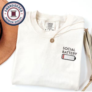 May include: Cream-colored t-shirt with the embroidered text "SOCIAL BATTERY" and a low battery graphic. A gold necklace with a pendant rests on the shirt. The shirt has a tag that says "COMFORT COLORS". A circular embroidered patch is in the upper left corner.