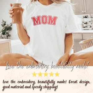May include: White t-shirt with the word "MOM" in red block letters. The shirt is being worn by a person. The image also includes a glass of iced coffee with a straw.