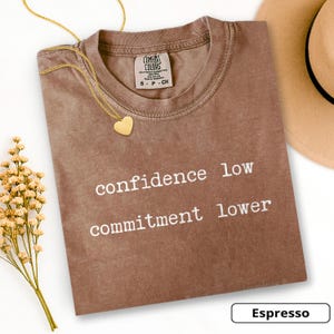 May include: Espresso-colored t-shirt with the text "confidence low commitment lower" in white. A gold heart pendant necklace and a tan hat are also visible. The shirt has a Comfort Colors tag.